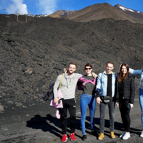 Cinco pessoas em pé em frente a uma paisagem rochosa e vulcânica com uma montanha e manchas esparsas de neve ao fundo.