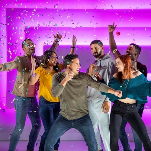 A group of six people partying with confetti against a pink and purple lit background, smiling and dancing.