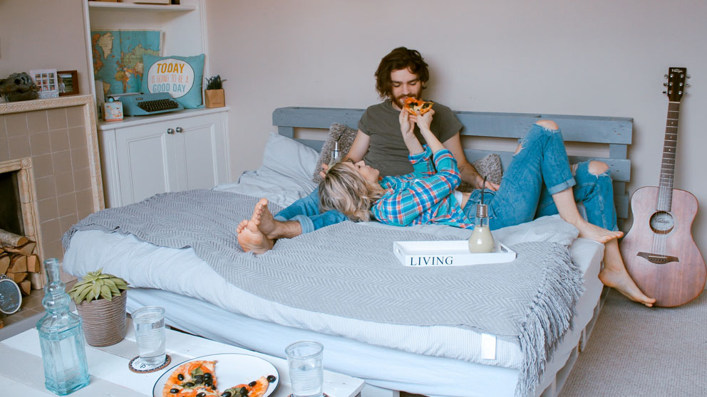 Iemand zit op een bed in een knusse kamer, omringd door kussens en persoonlijke spullen, met een roze gitaar in de buurt.