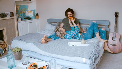 A person sits on a bed in a cozy room, surrounded by pillows and personal items, with a pink guitar nearby.