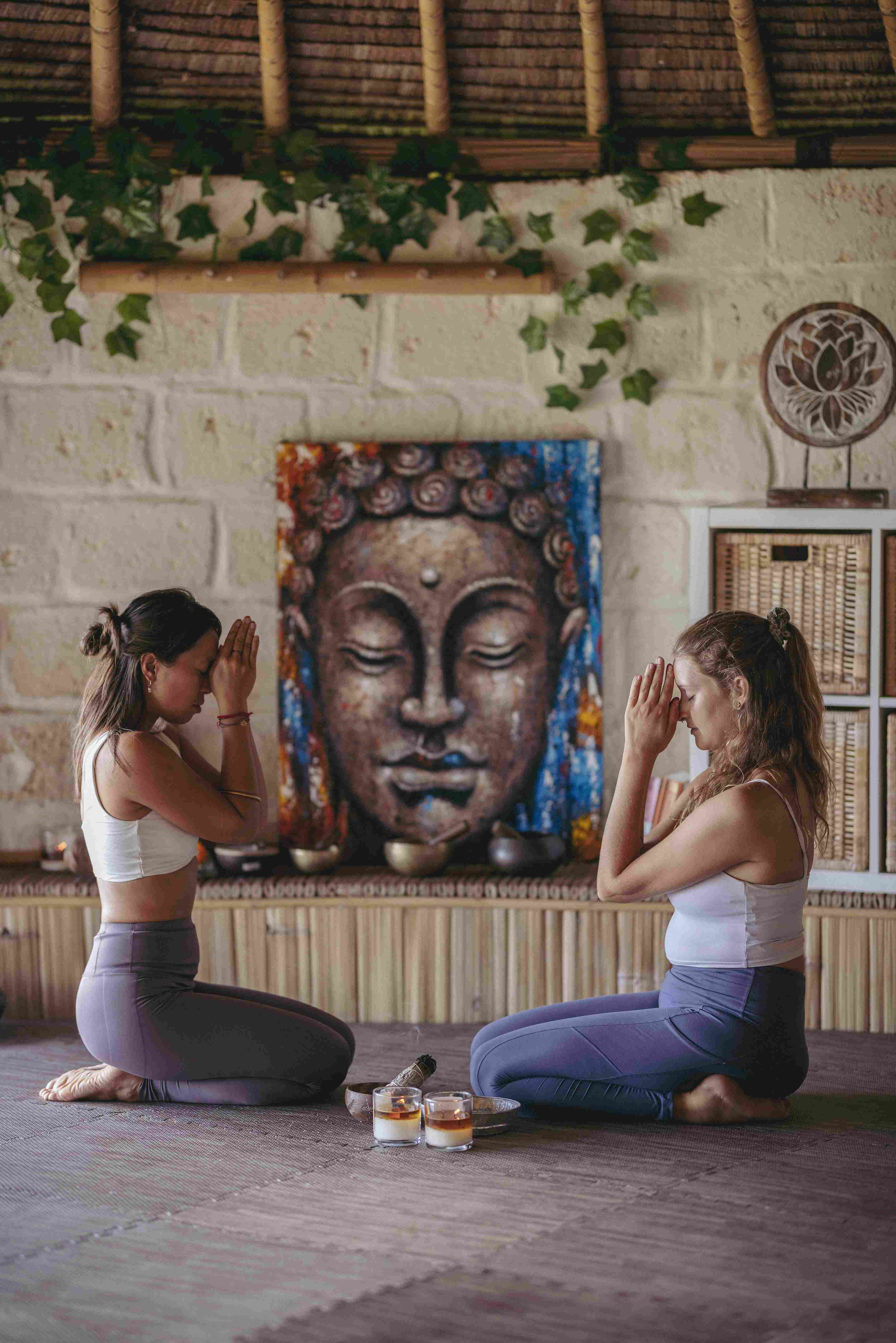 Two students practicing partner meditation in the bamboo yoga shala with a large Buddha painting background.