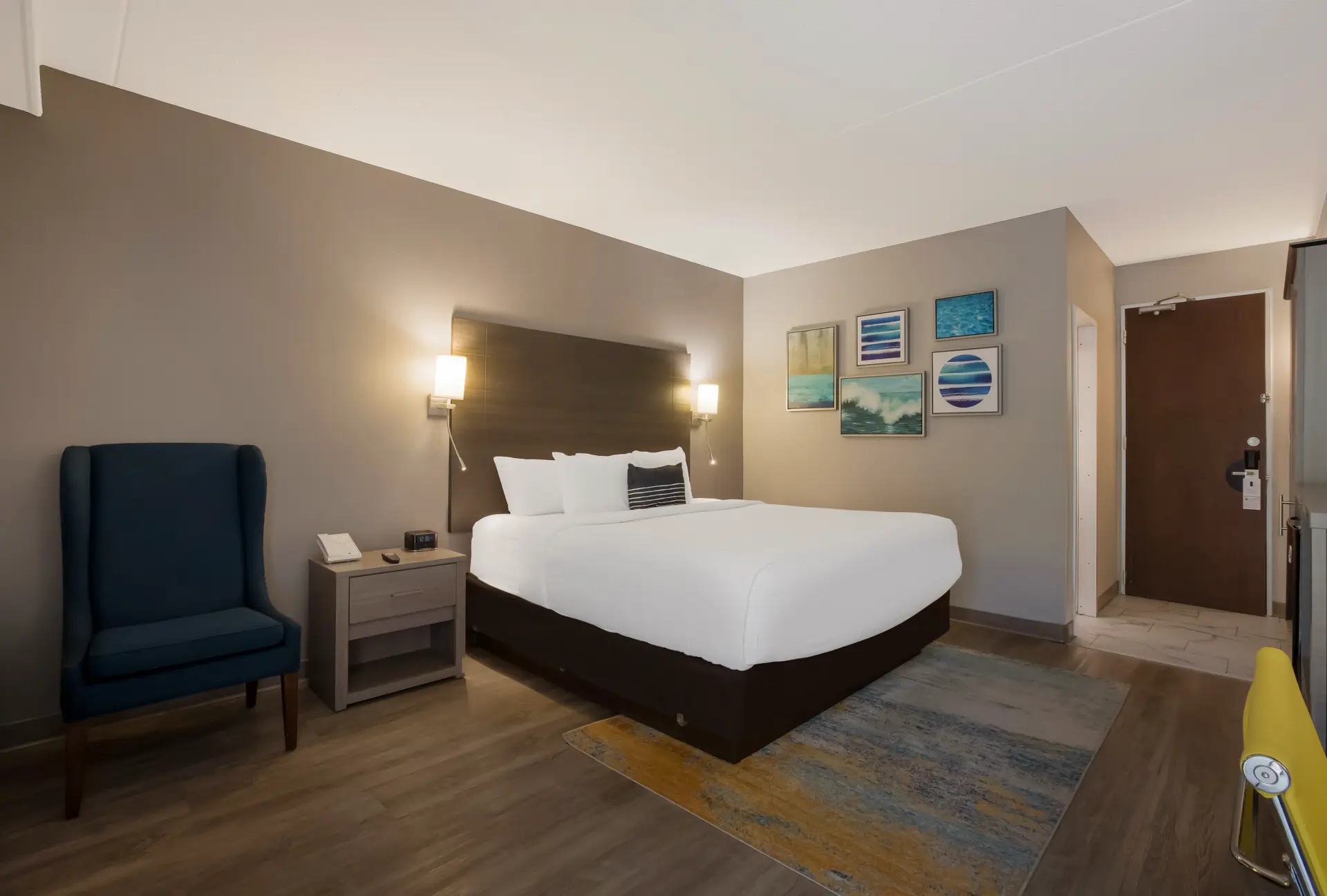 Hotel room with bed, nightstand, chair, and paintings on the wall.