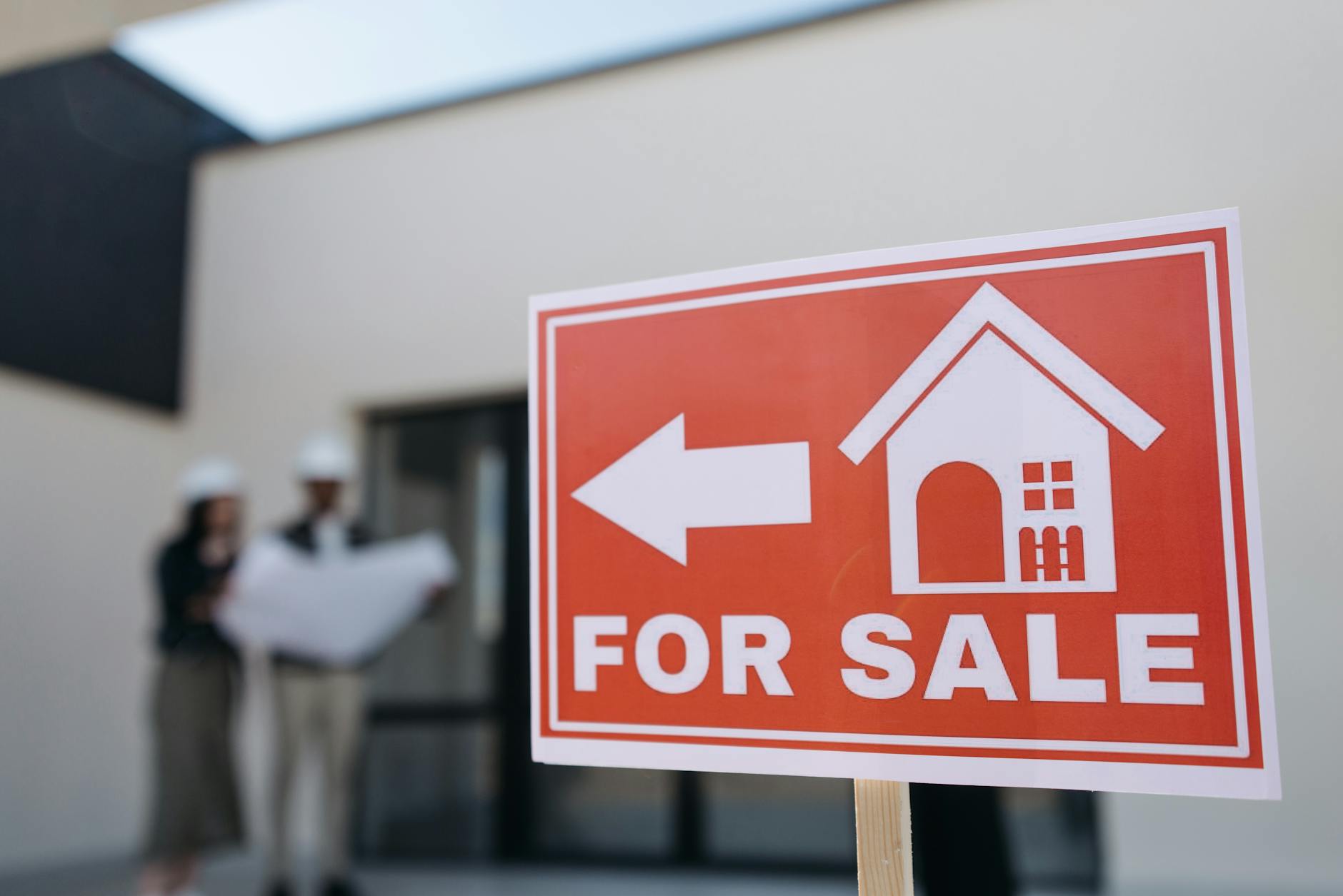 Red For Sale sign with house icon and arrow in front of a building where two architects review blueprints