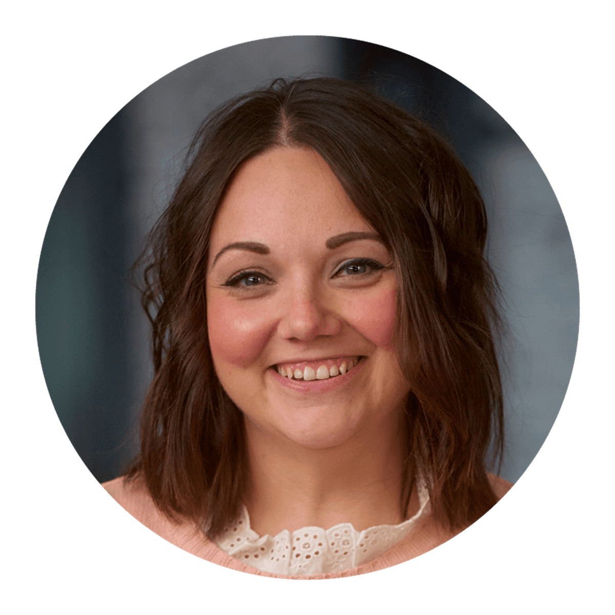 A circular studio‑style portrait of a person with shoulder‑length brown hair, wearing a light pink top with a decorative neckline, set against a softly lit background.