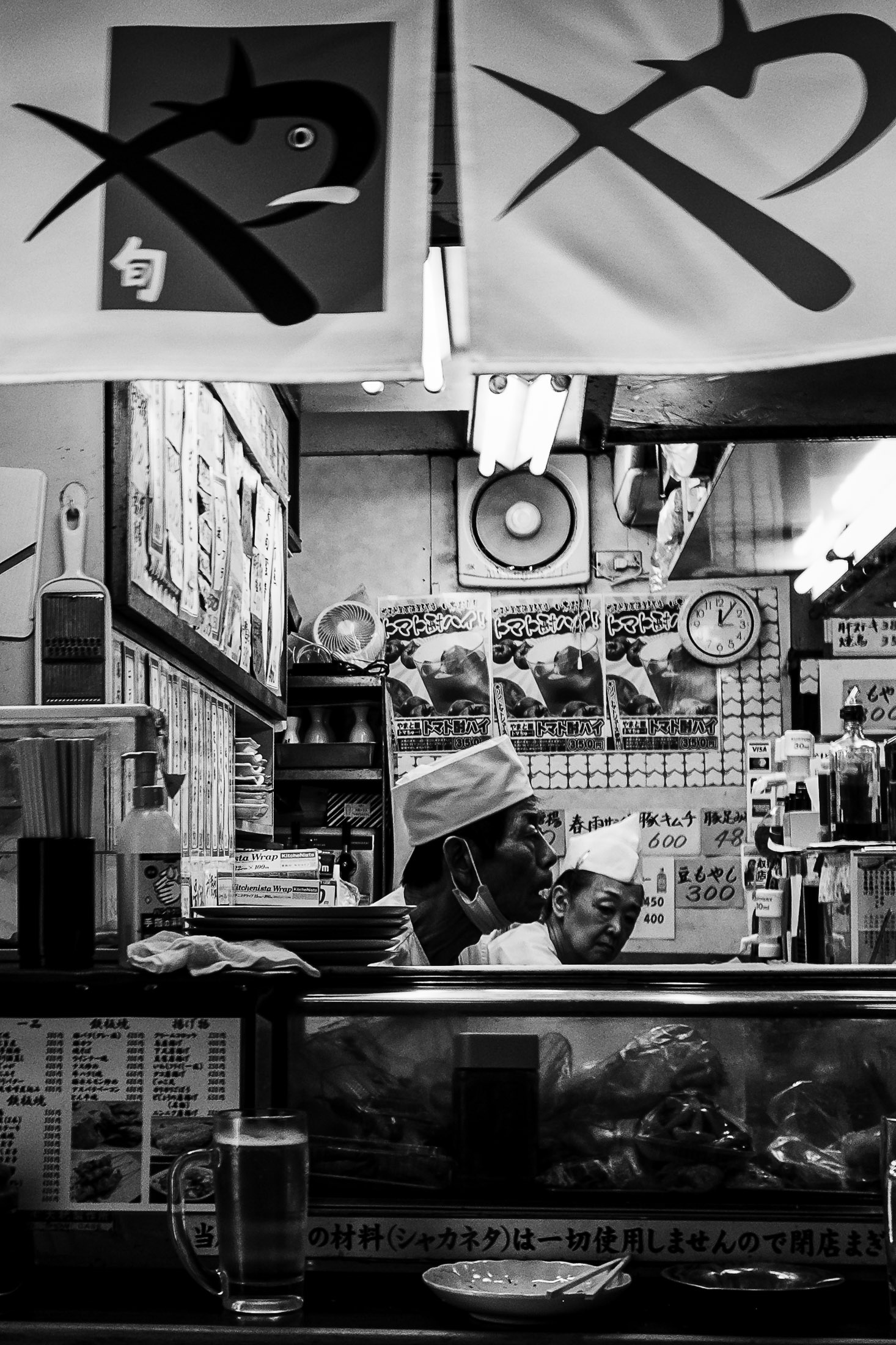 Black and white photograph of chefs working inside a small Japanese restaurant kitchen, street photography by Richard Peterson.