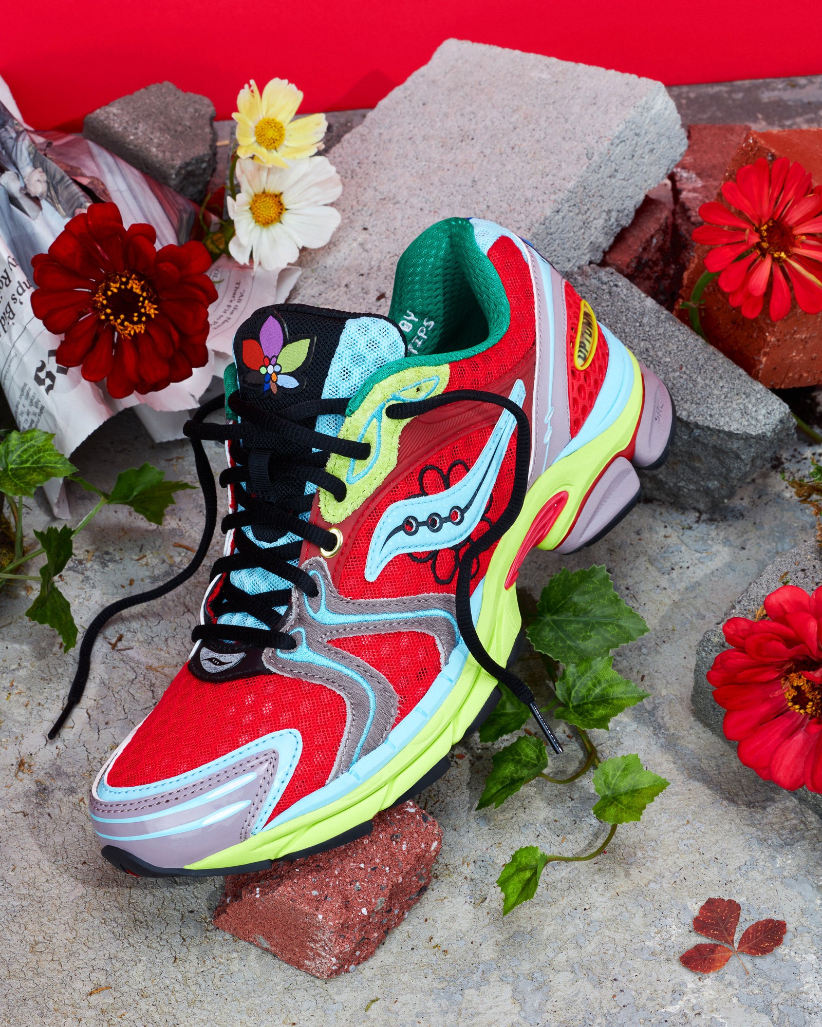 Red Jae Tips x Saucony sneaker styled on stone with flowers and scattered brick pieces against a bright backdrop.