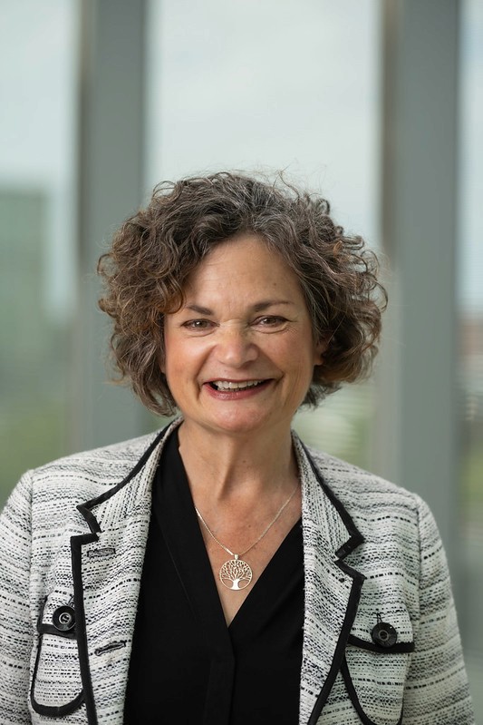 A smiling woman with curly hair, wearing a black top and a patterned blazer, poses in front of a window.