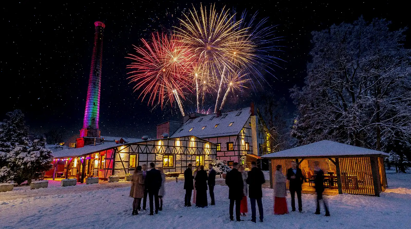 Sylwester 2025/2026 w Wrzosowym Tartaku — zimowa sceneria, pokaz fajerwerków nad oświetlonym budynkiem i grupą gości.