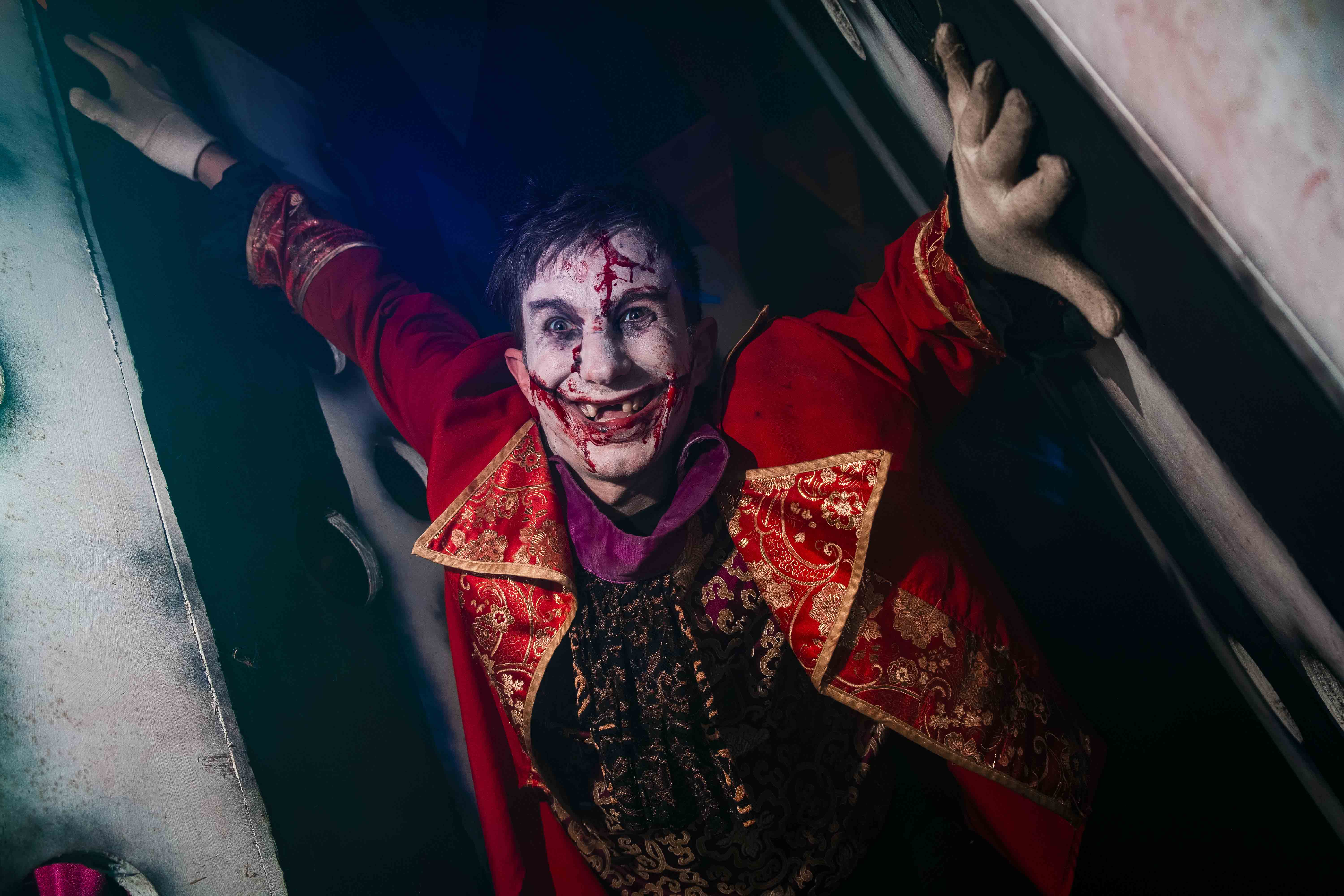 A person dressed as a sinister clown with white face paint, red accents, and smeared makeup leans menacingly against a wall in a dimly lit setting, creating a chilling, haunted atmosphere.