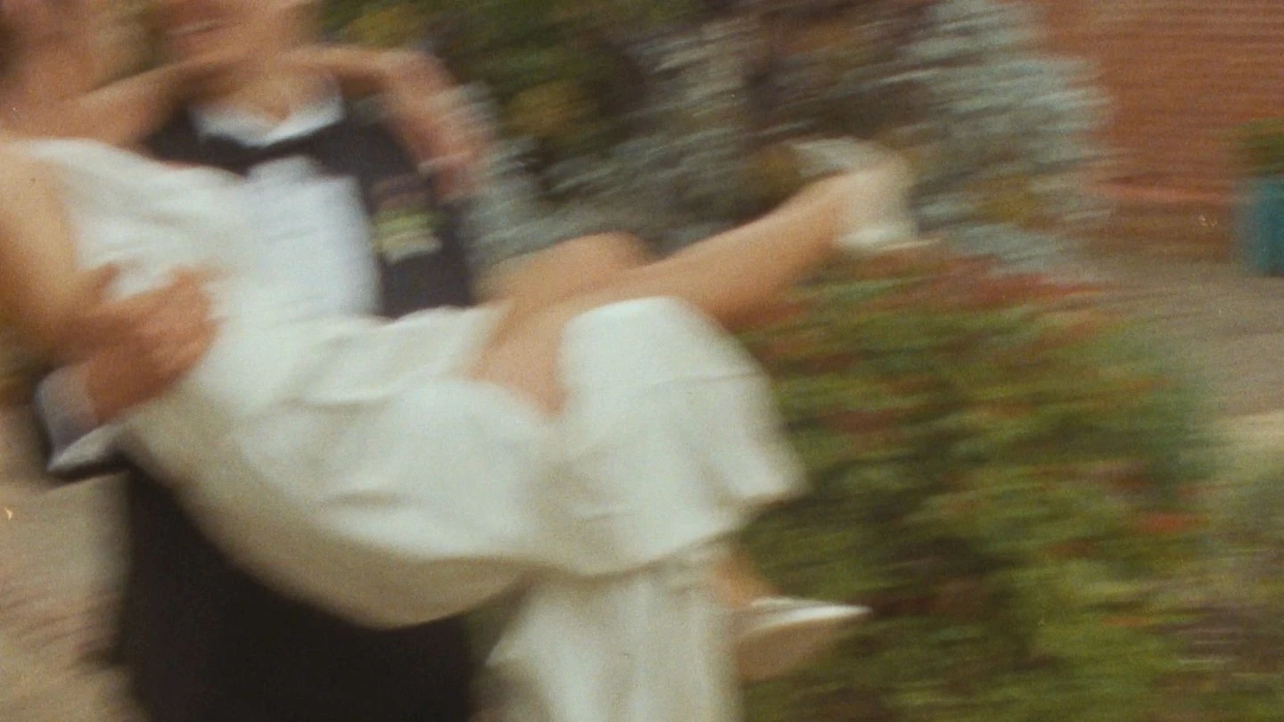 A blurry photo captures a person in a black tuxedo joyfully carrying another person in a flowing white dress, exuding a celebratory and romantic atmosphere.