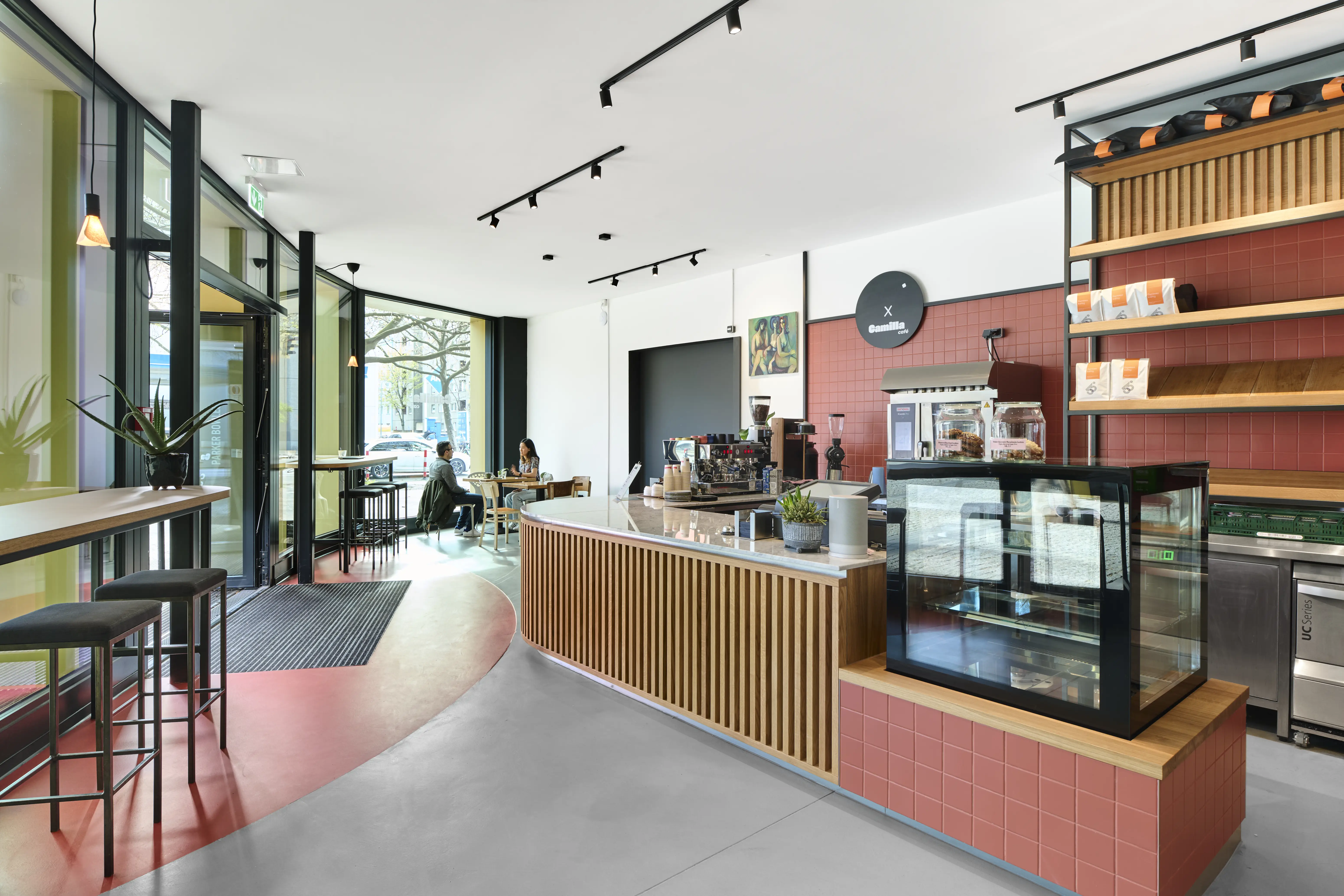 Overview of CAFÉ CAMILLA with a curved wooden vertical-slat counter with white marble top, terracotta-red tiled walls, a red floor circle and a round black Camilla logo disc.