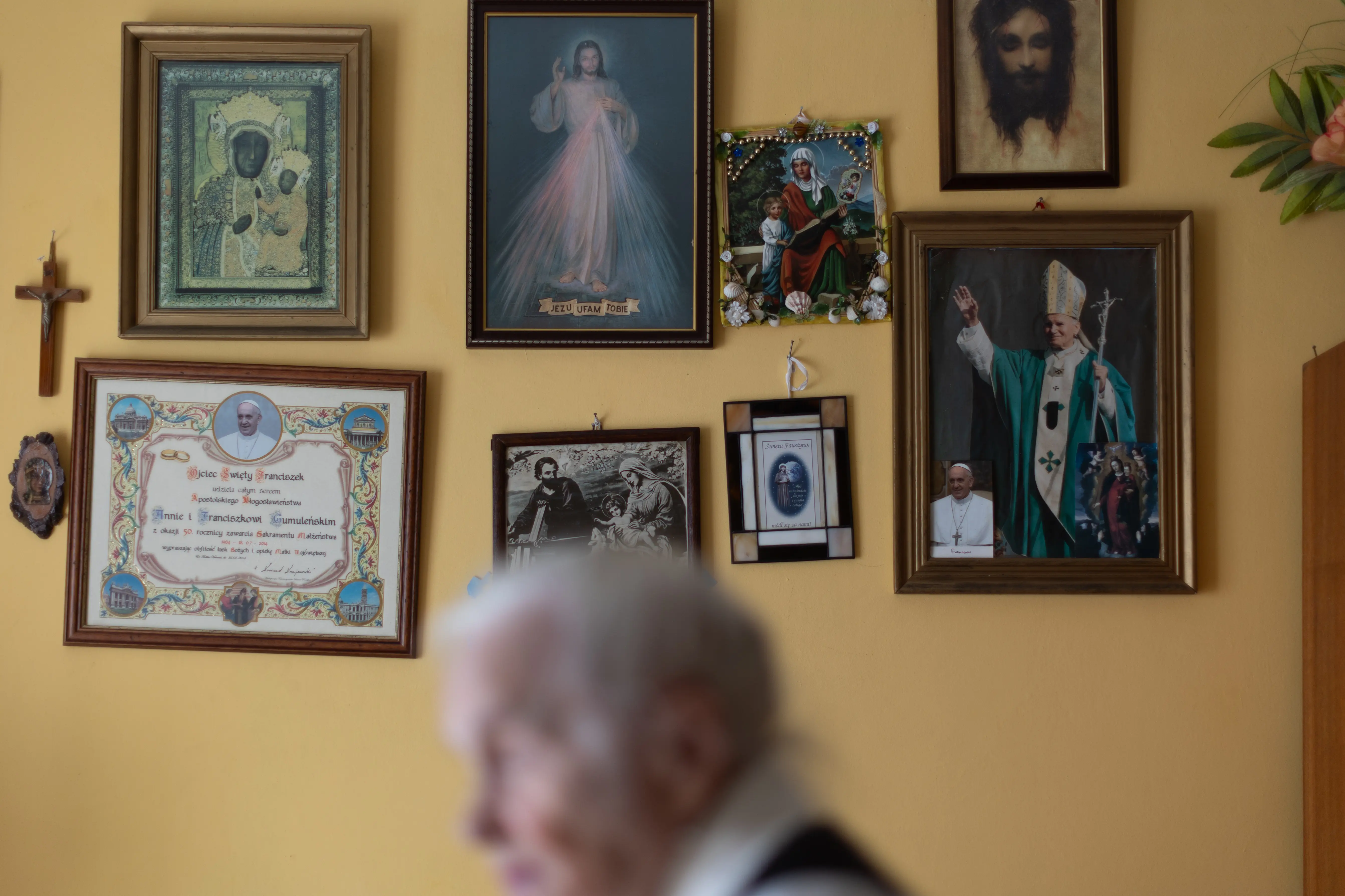 Sylwetka starszej, siwek kobiety rozmuta na tle żółtej ściany z wieloma religijnymi obrazami.