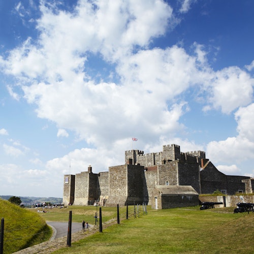 ปราสาทหินขนาดใหญ่ที่มีป้อมปราการภายใต้ท้องฟ้าที่มีเมฆบางส่วน ล้อมรอบด้วยเนินเขาที่ปกคลุมด้วยหญ้า มีผู้คนอยู่ในระยะไกล