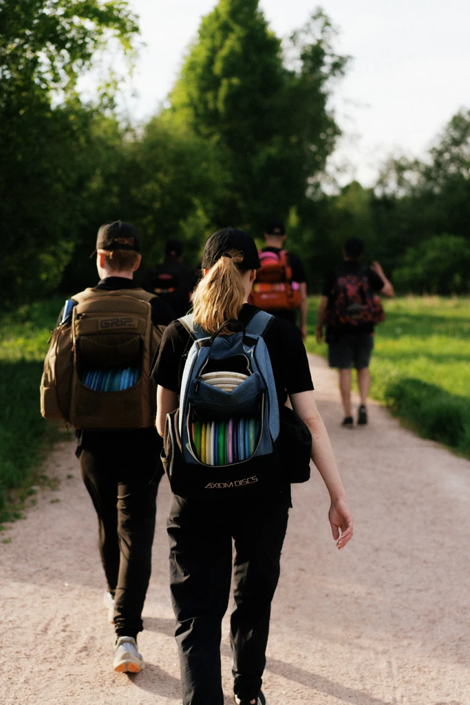 A group of disc golfers walks along a scenic dirt path surrounded by lush greenery, heading toward the next hole. They are wearing casual sportswear and carrying disc golf backpacks loaded with colorful discs. The two players in the foreground, one male and one female, have backpacks branded with "Grip" and "Axiom Discs," emphasizing their passion for the sport. The warm sunlight and natural setting highlight the camaraderie and shared adventure of playing disc golf together.