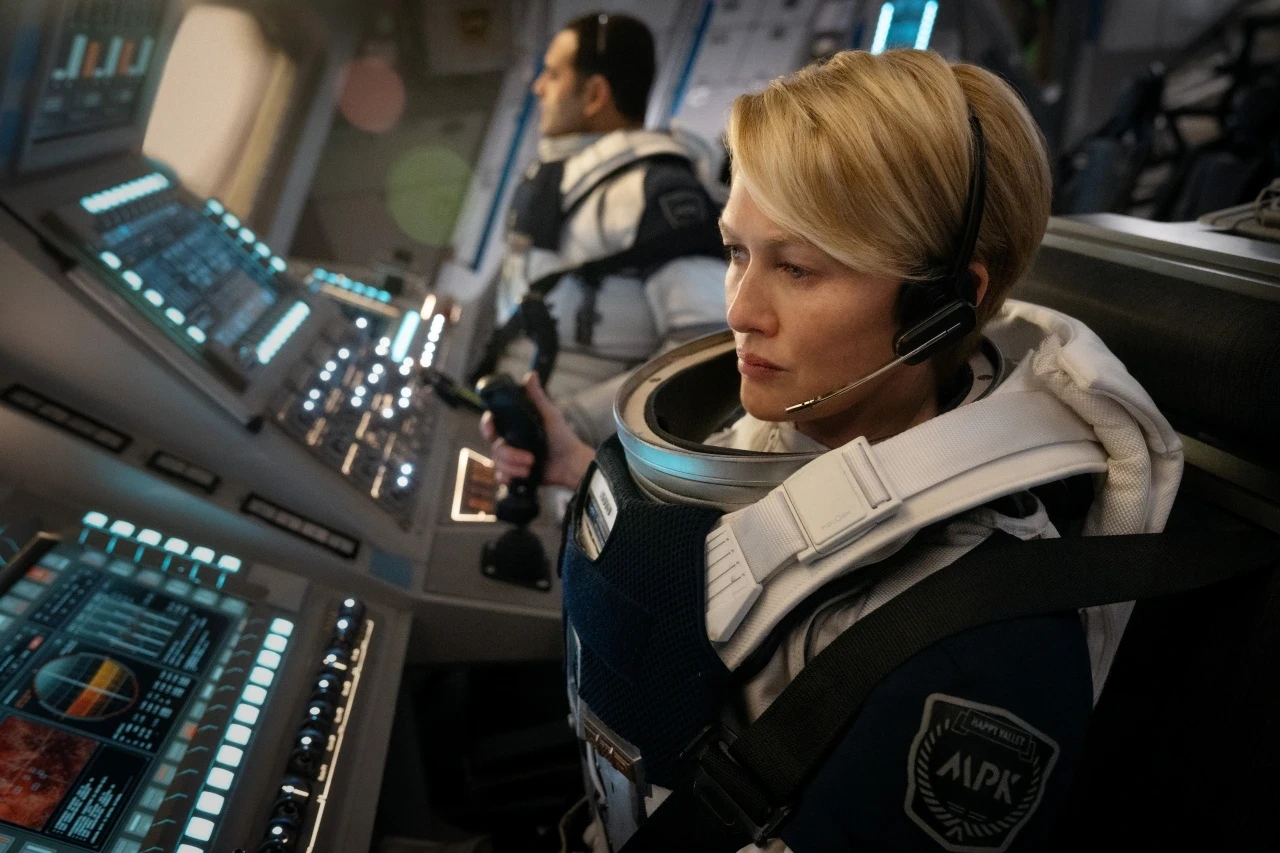 Two astronauts wearing space suits and headsets are inside a spacecraft, focused on operating advanced control panels with illuminated screens.