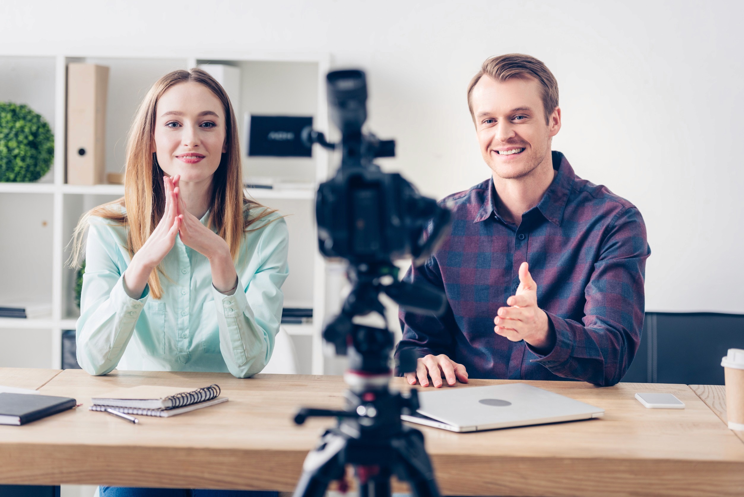 Two professional influencers, a woman and a man, filming content at a desk in a modern office setting. The woman, wearing a light blue shirt, gestures thoughtfully, while the man in a checkered shirt smiles and gestures towards the camera. A camera on a tripod is set up in front of them, capturing their engaging and confident presentation. They have a laptop, notebook, and coffee cup on the desk, emphasizing their professional and knowledgeable approach.