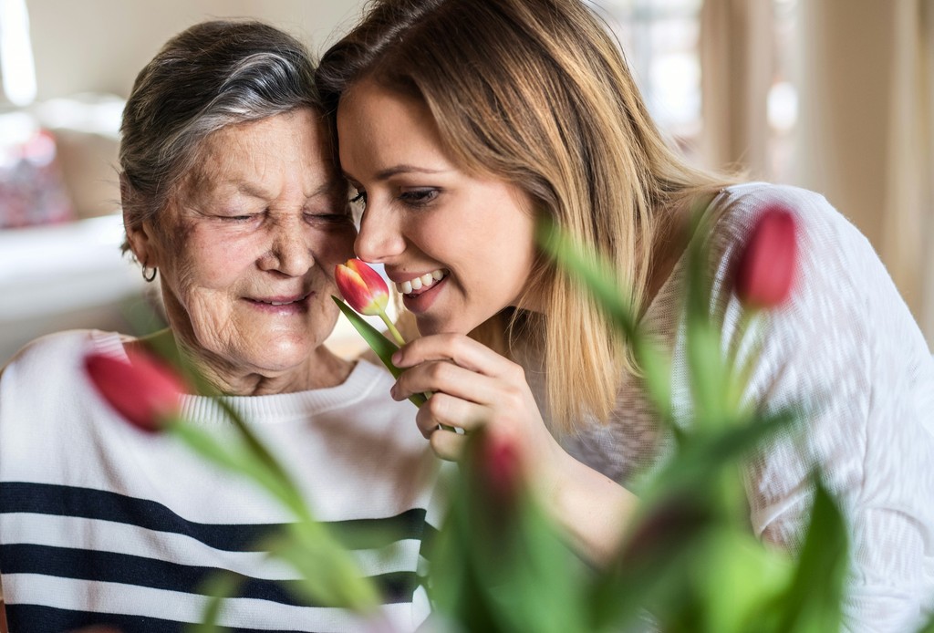 A joyful elderly woman and her granddaughter share a moment of closeness, enjoying the scent of a tulip together in a warmly lit room adorned with vibrant flowers.