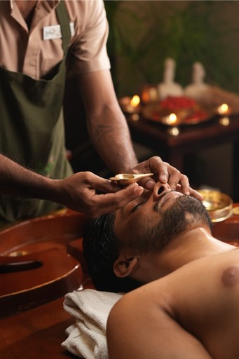 Ayurvedic body purification and detox therapy – A man receiving a synchronized Pinda Sweda (herbal pouch massage) from two therapists in a traditional wellness spa setting.