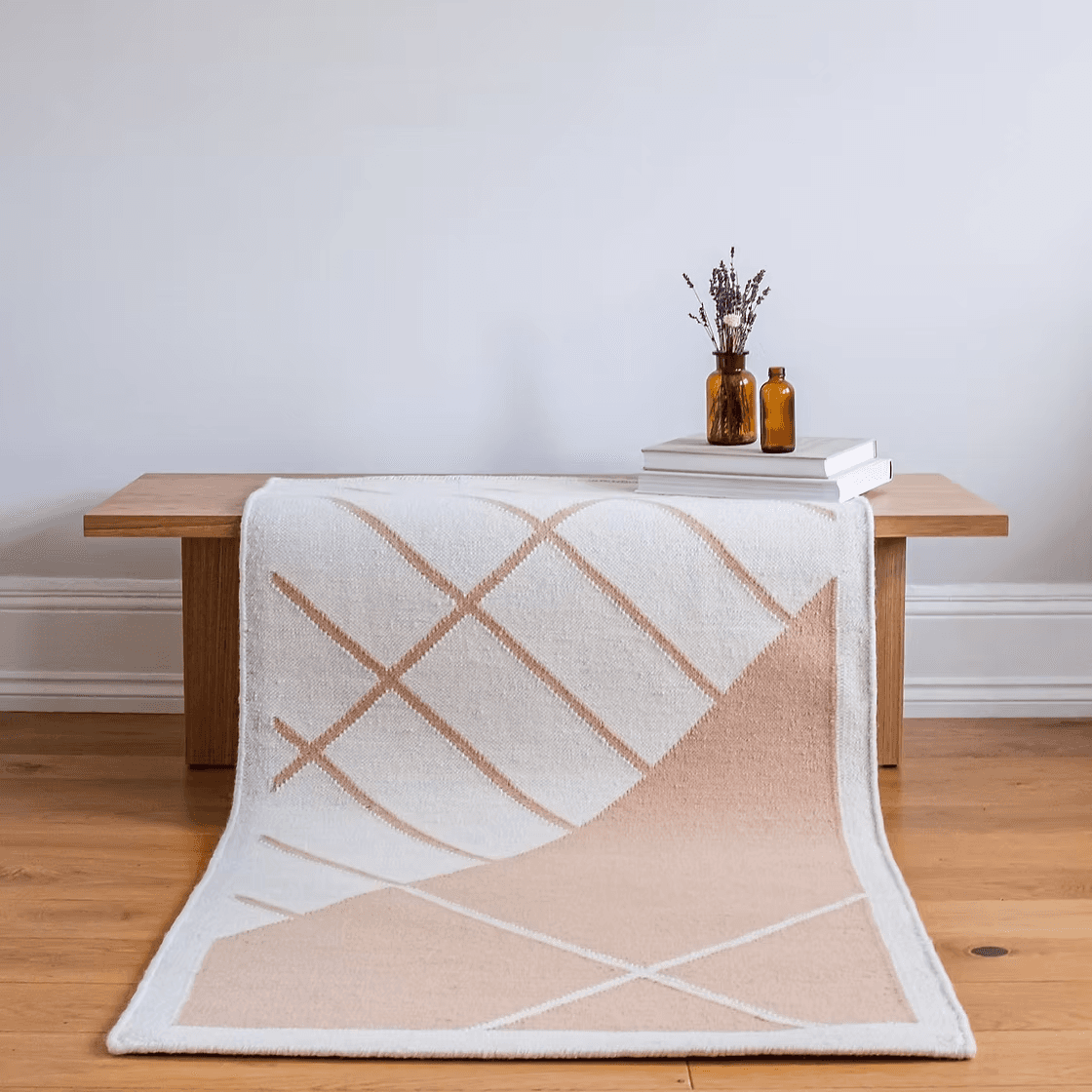 A minimalist wooden bench is adorned with a light-colored geometric patterned rug draped over it, accompanied by amber glass vases and stacked books, set against a clean, white wall background.