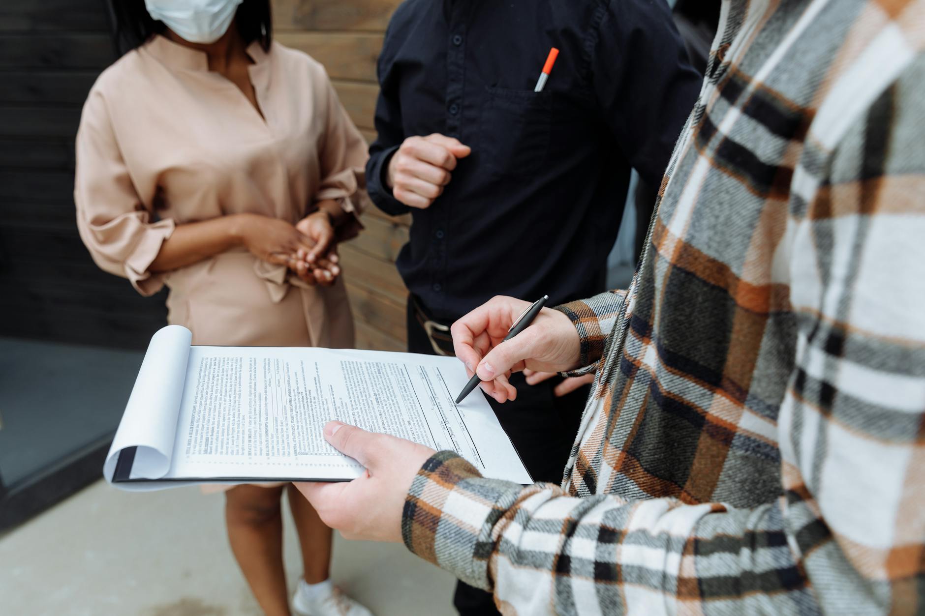 Person in plaid shirt reviewing a printed contract document with two people outdoors, one wearing a face mask