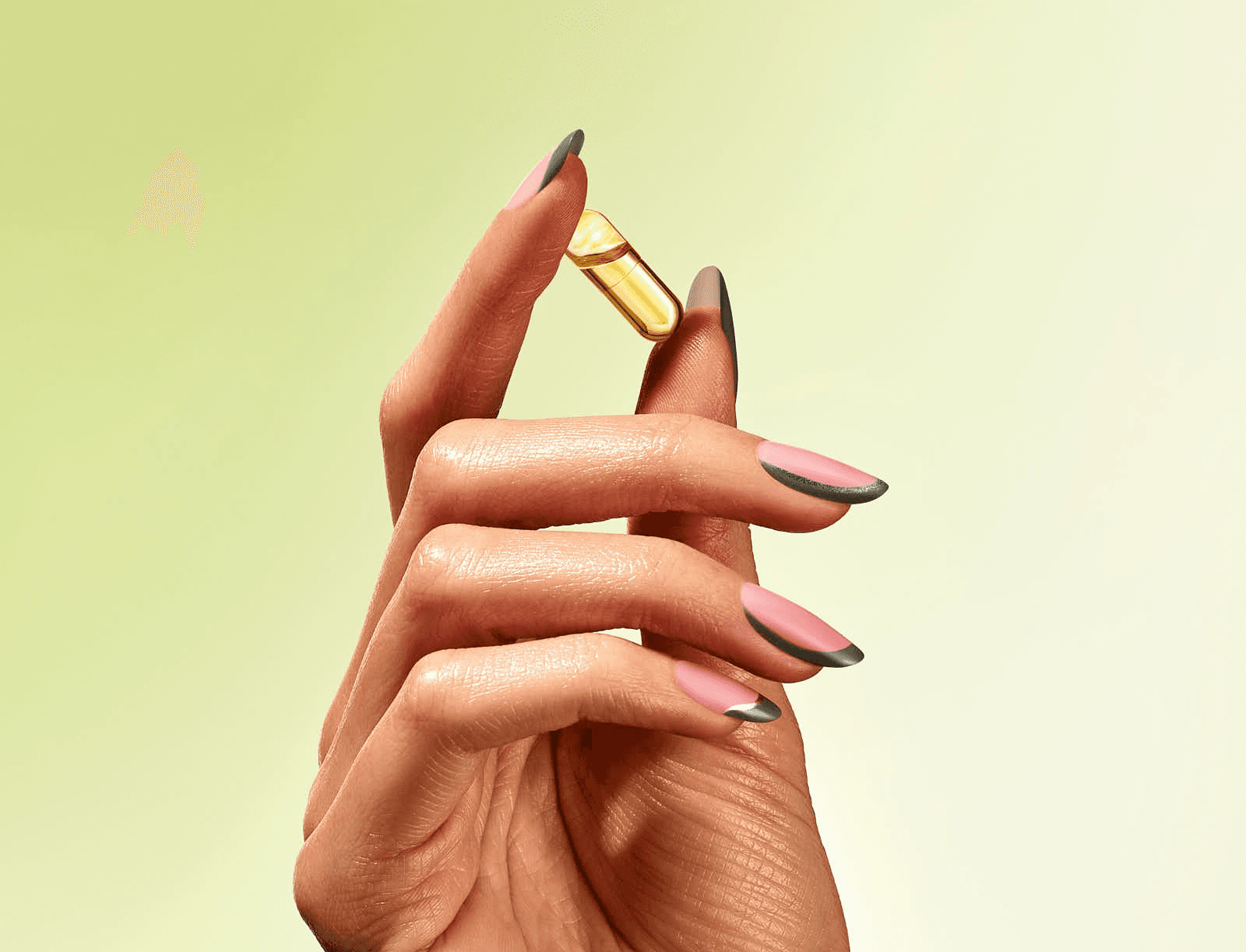 Hand holding a vitamin capsule with stylish pink and gray nails on a light green background.