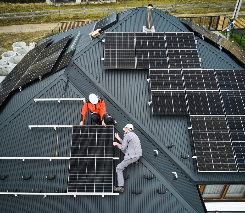 Luftaufnahme von zwei Arbeitern, die schwarze Solarmodule auf einem geneigten Wohnhausdach installieren, um erneuerbare Energien und nachhaltige Lösungen für Wohngebäude zu fördern.
