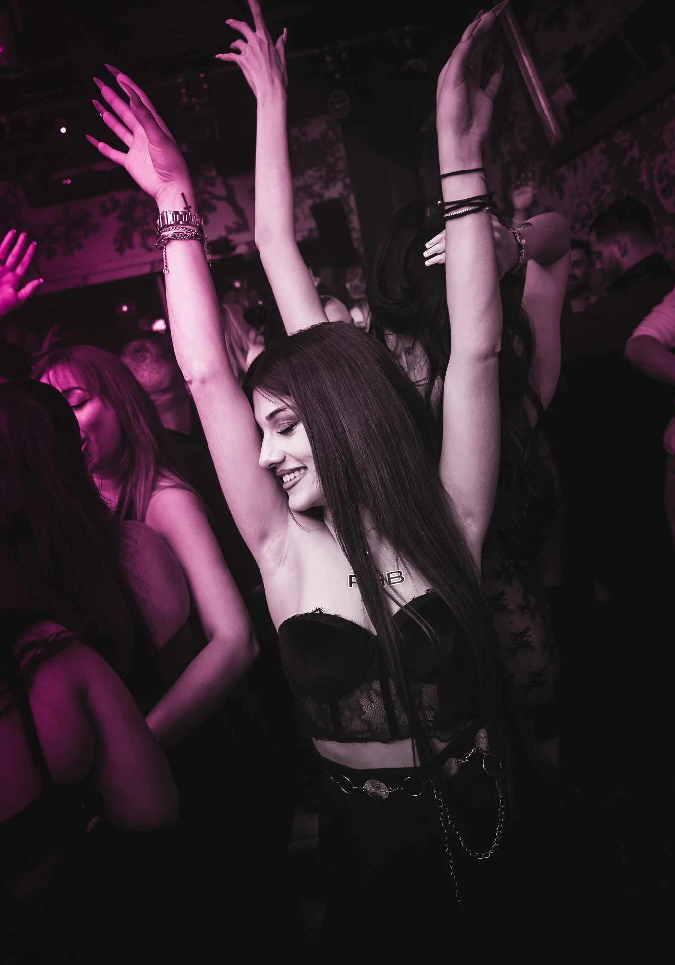 A person at the RHB party smiles and laughs with their arms raised high in celebration. They are in a crowd and wearing a black lace crop top under pink lighting.
