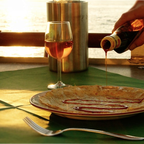 一个人在盘子里的可丽饼上倒糖浆，旁边是一杯葡萄酒，背景是日落景色。