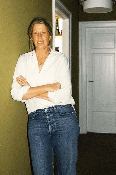 A casually dressed Alex Degel with her arms crossed, wearing a white shirt and blue jeans, standing against a softly lit background.