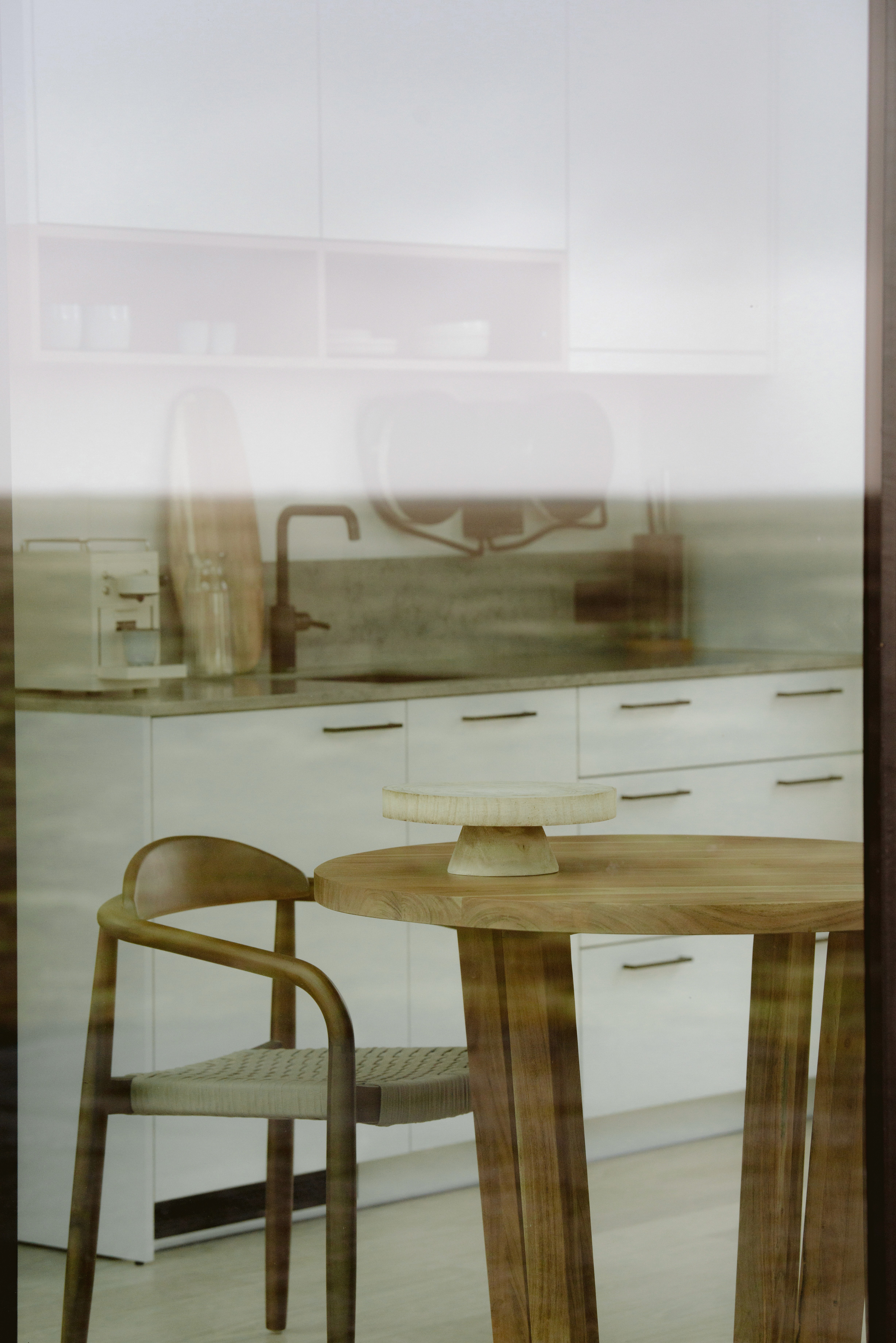 Modern kitchen and dining area with minimalist white cabinetry, wooden dining table and chair, viewed through floor-to-ceiling glass in a contemporary design-led cabin overlooking an open landscape.