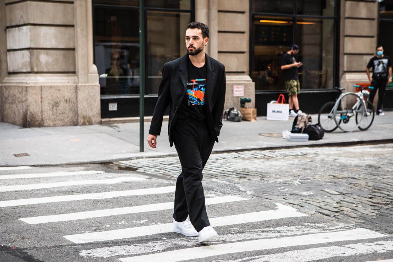 Street-style portrait of a Eric Whiteback walking across a crosswalk in a black blazer, graphic tee, and white sneakers.