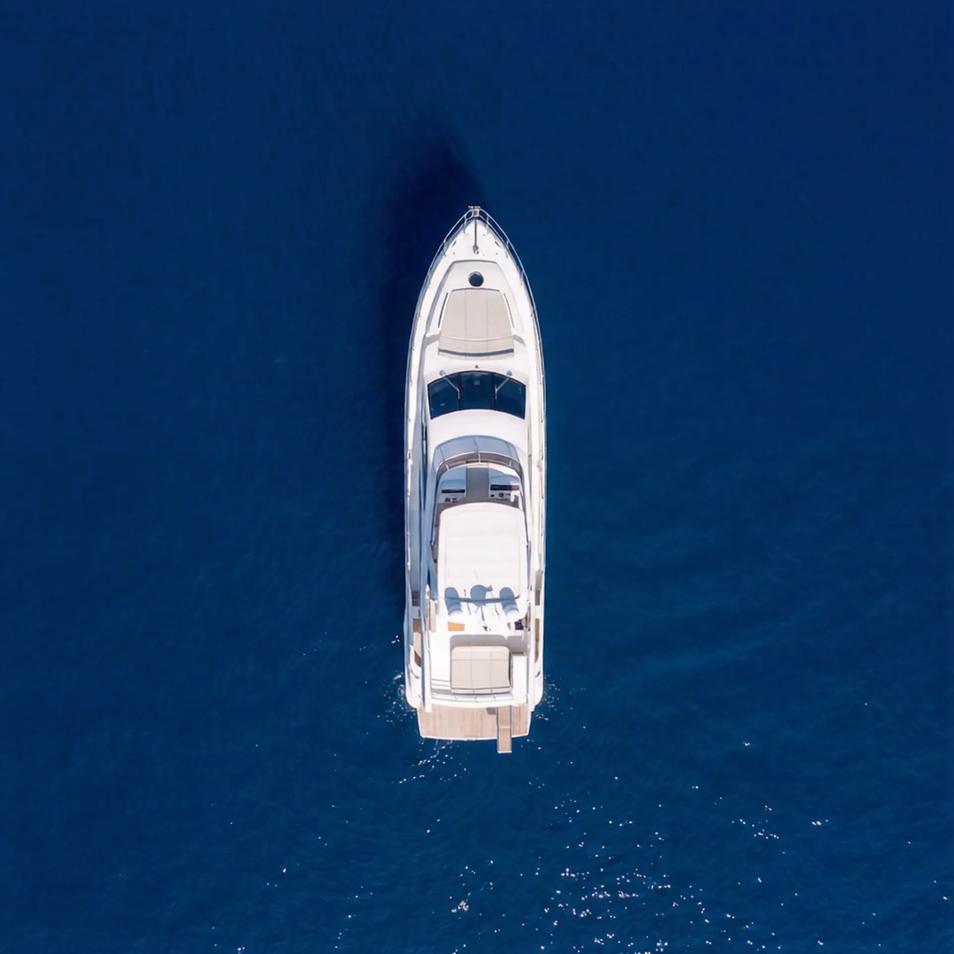 Aerial top-down view of an Azimut 68 luxury motor yacht underway on deep navy blue open ocean water, showing the full EcoDeck composite deck layout across the flybridge, cockpit, and aft swim platform with cream sunpad cushions and teak-finish stern