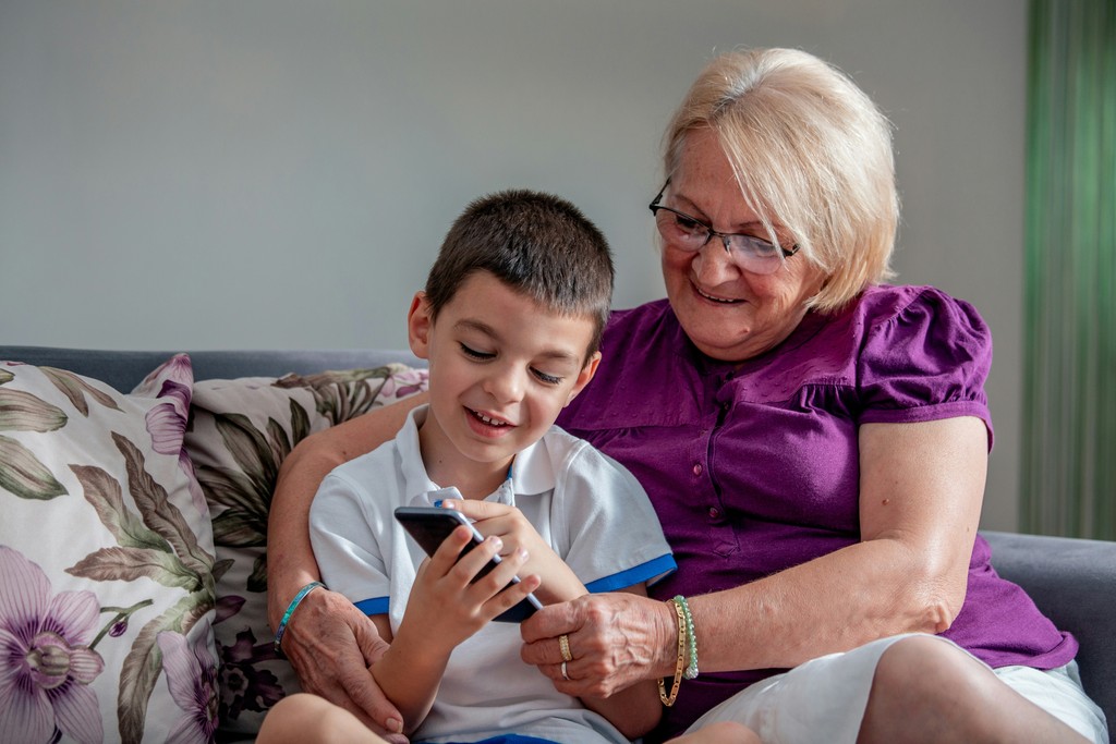 A grandmother and her young grandson share a tender moment on the couch, smiling as they look at a smartphone together. The scene captures the joy of generational bonding and the shared curiosity and learning between them, highlighting the warmth and love in their relationship.