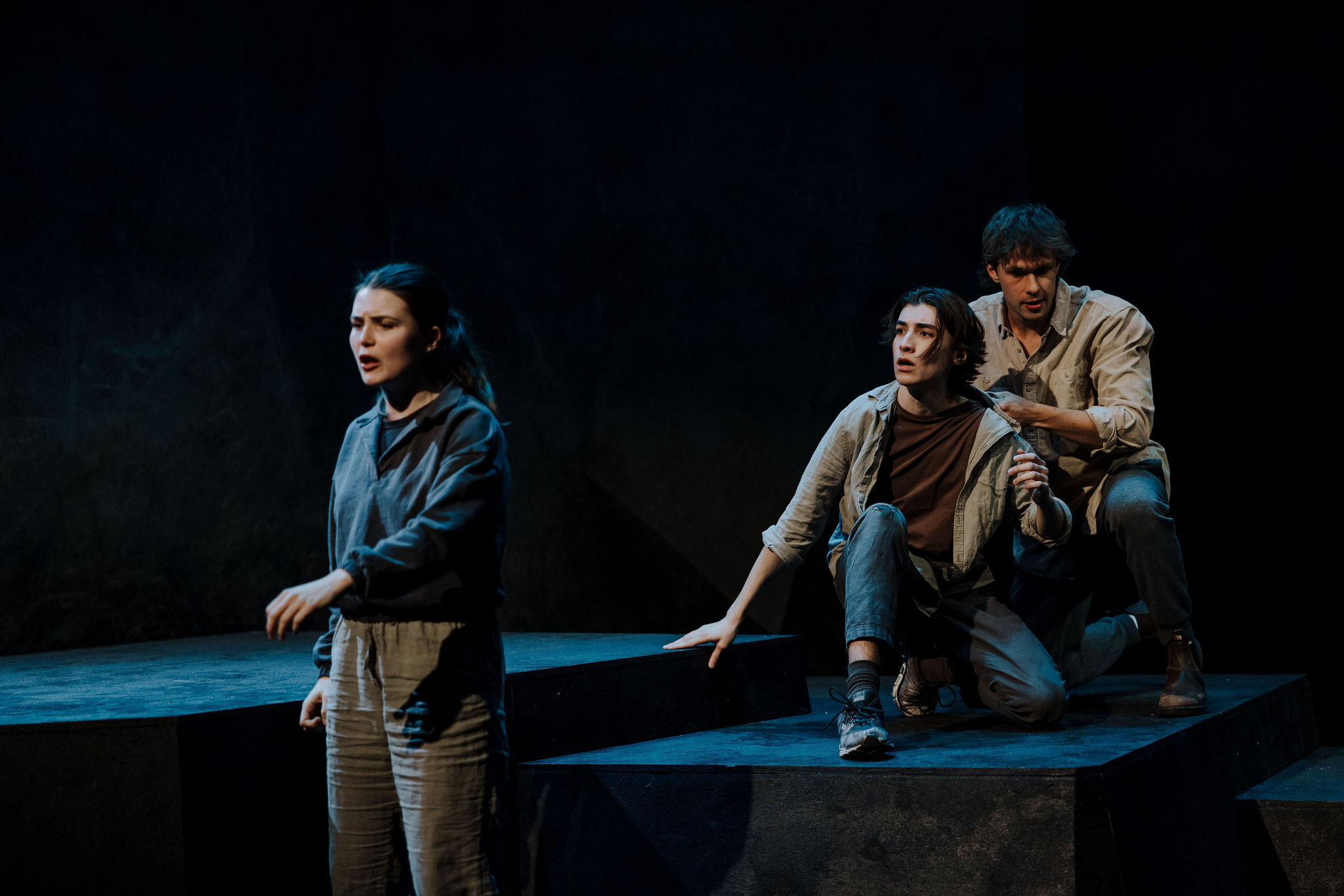 A performer sings or speaks in the foreground while two others crouch behind them on a stepped platform, all wearing muted casual clothing.