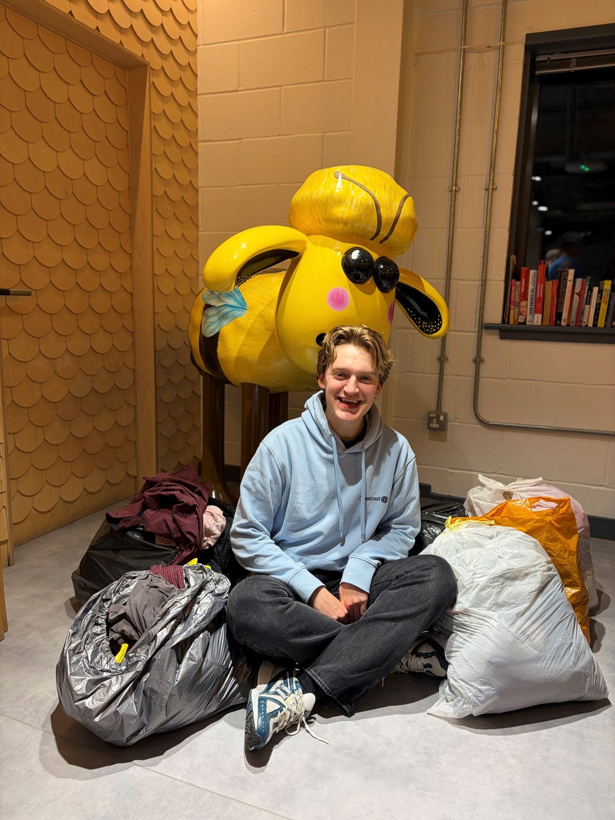 Person sitting on the floor with several large bags of clothes, in front of a bright yellow bee sculpture indoors.