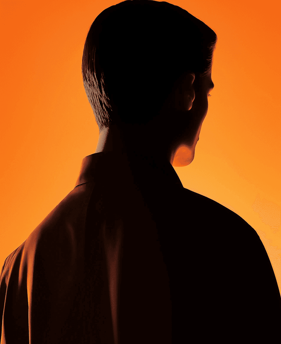 A man in a black suit stands confidently against a vibrant orange background.