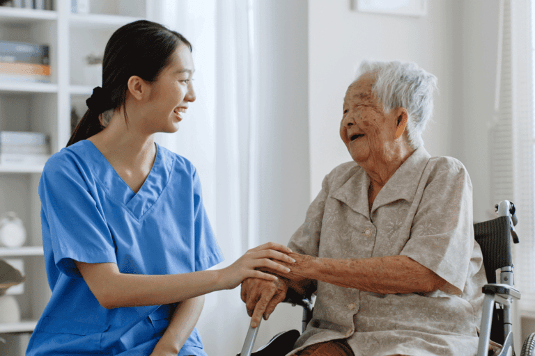 A care assistant in blue scrubs warmly interacting with and assisting an elderly woman, showing compassionate senior care
