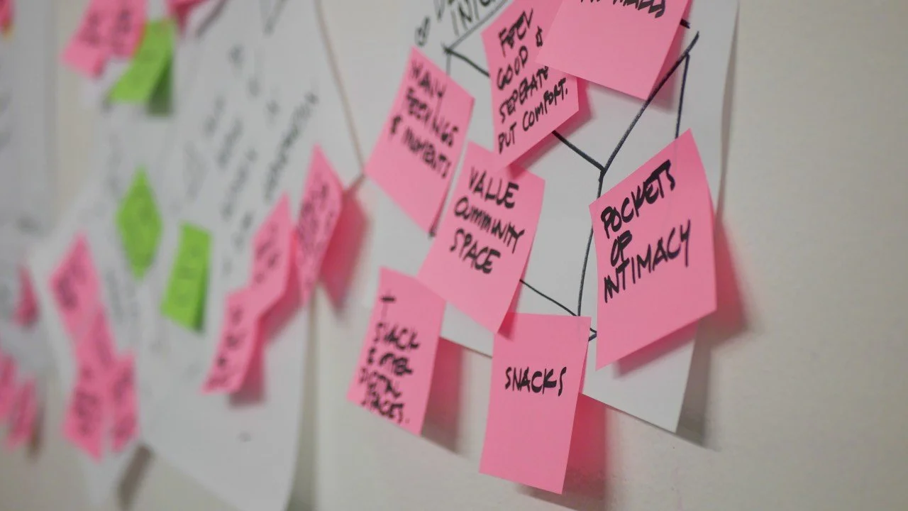 Close-up of colorful sticky notes on a wall, with pink and green notes displaying various handwritten tasks and ideas.