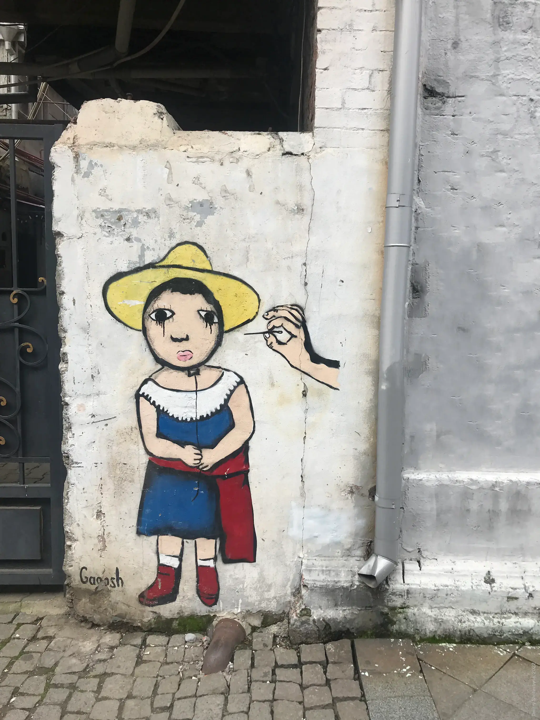 A small stencil-style mural on a worn white brick wall depicts a sad-eyed child wearing a yellow hat, a blue dress, and red shoes. Her disembodied right hand reaches toward the viewer.
