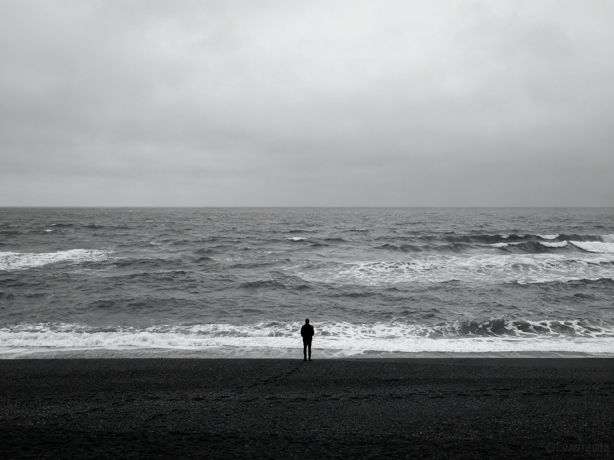 An editorial photograph of a solitary figure standing at the edge of a vast beach during an overcast