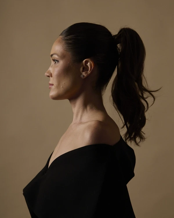 A young person with curly hair smiles at the camera while sitting in front of a laptop against a dark background.
