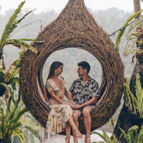 一對情侶坐在一個大型淚滴形的柳條巢中,懸掛在熱帶植物之間,互相凝視並握著手。