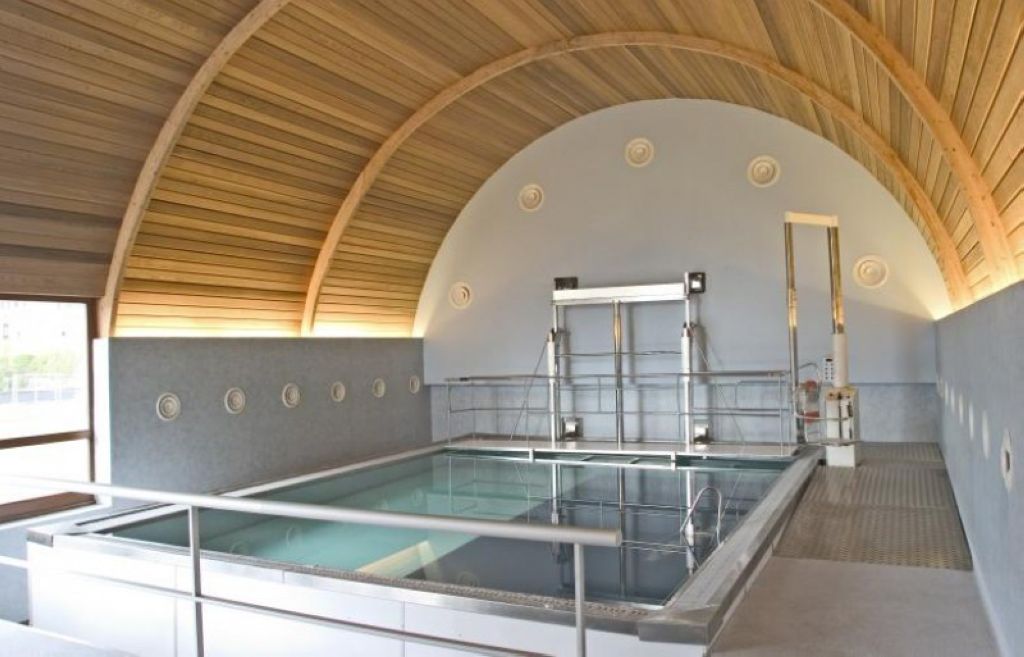 Indoor hydrotherapy pool with warm-toned curved wooden arched ceiling, therapeutic water, metal safety railings, and treatment space