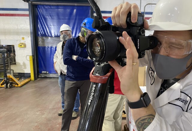 A person in safety gear uses a camera mounted on a tripod in an industrial setting, capturing visuals for an advertising agency Baltimore, while others in hard hats and masks stand in the background.