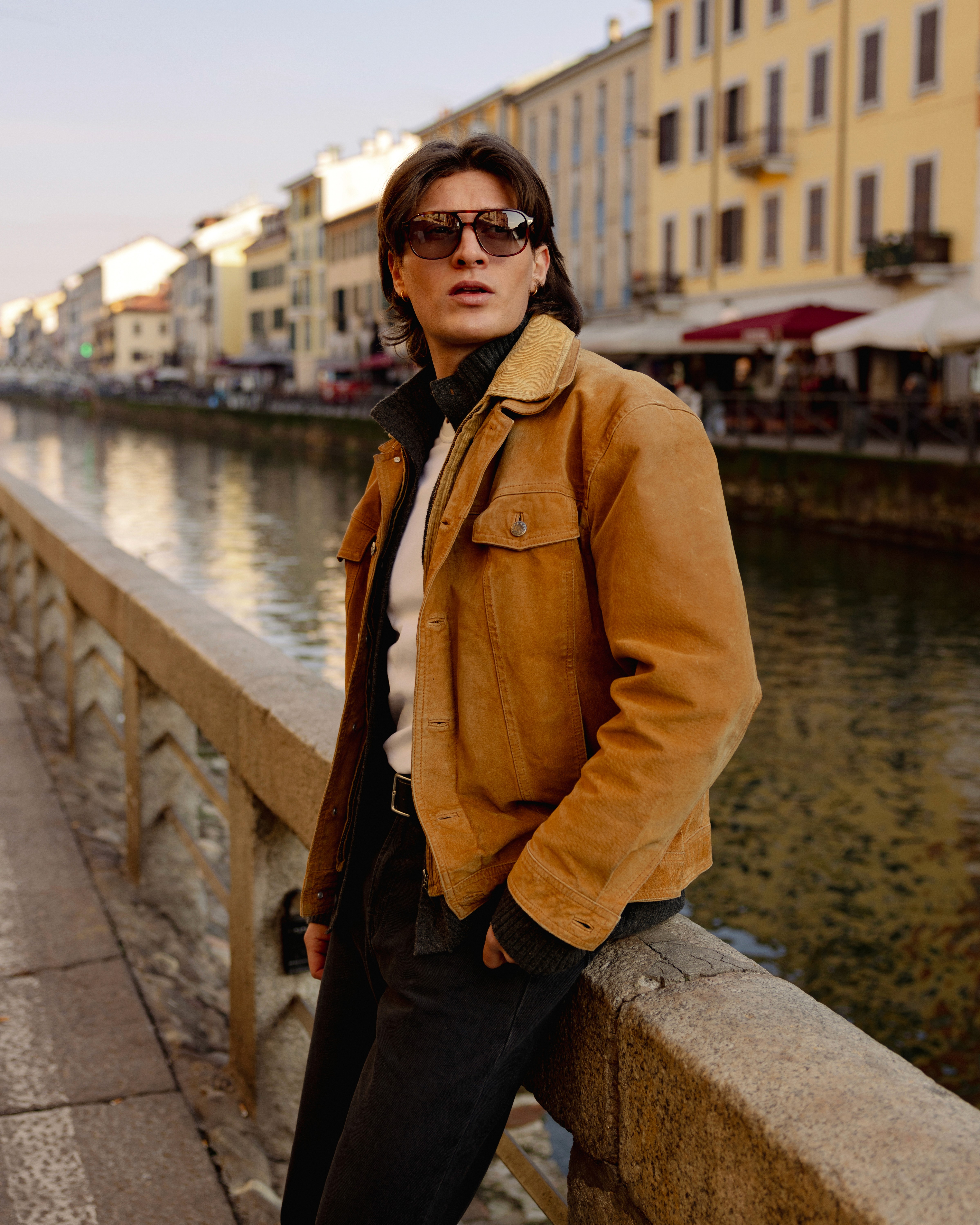 Un homme porte une veste cognac et des lunettes de soleil le long d'un canal
