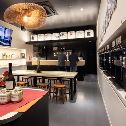 現代酒吧內部，三個人站著，長桌上有座椅、清酒瓶，以及牆上的一排分配器。