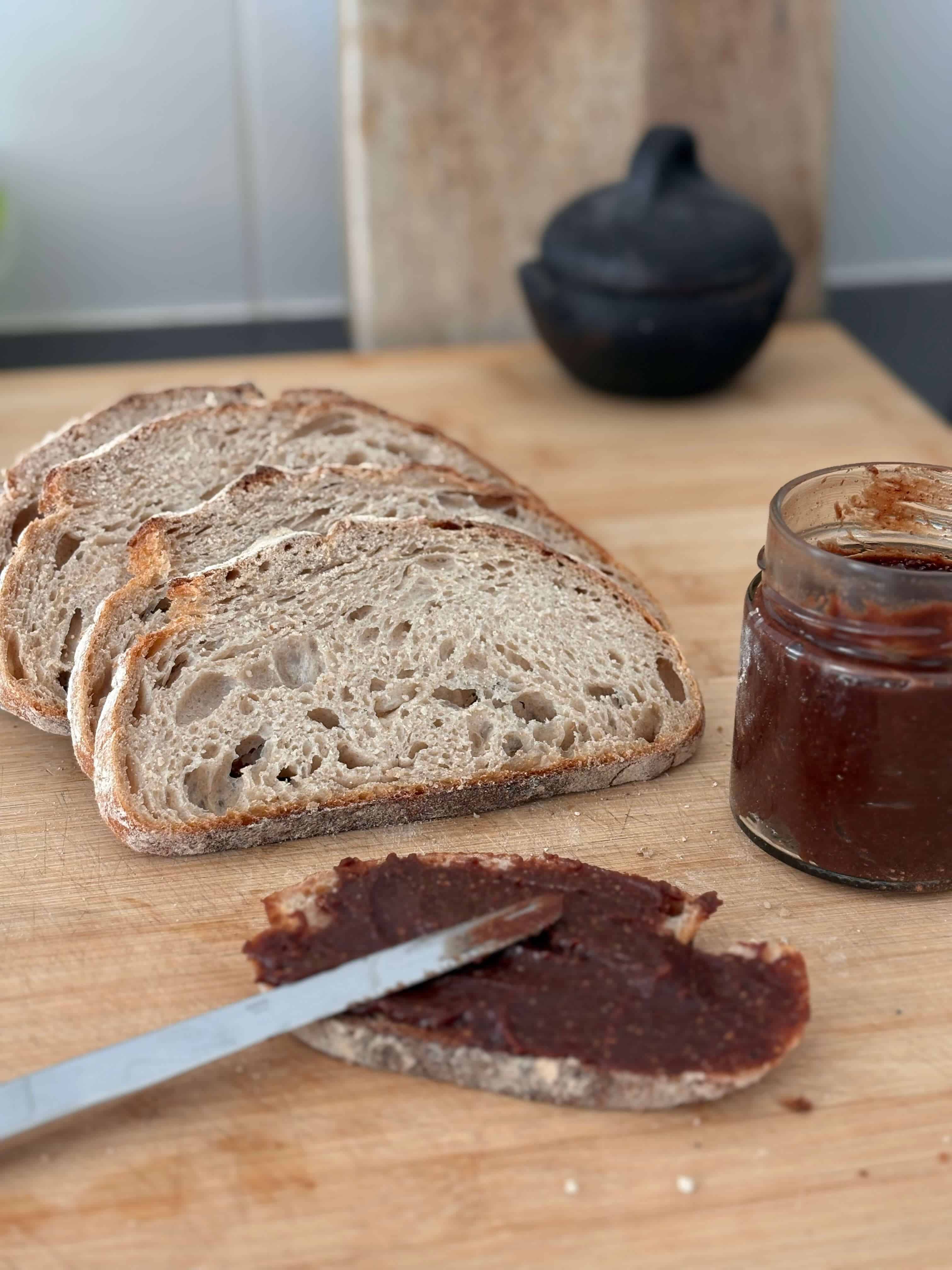 Homemade chocolate hazelnut spread being spread onto a slice of sourdough bread, with additional bread slices and a jar of spread on a wooden surface.