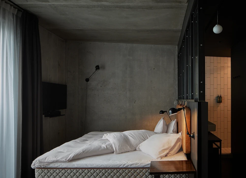 Bed beside concrete walls with open bathroom area in a Junior Suite.
