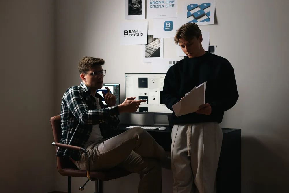 Felix Maier und Paul Schlager arbeiten im Büro an einem Webdesign-Projekt mit Computer und Designentwürfen der Marke „Base & Beyond“ – kreative Teamarbeit im Bereich Webdesign.