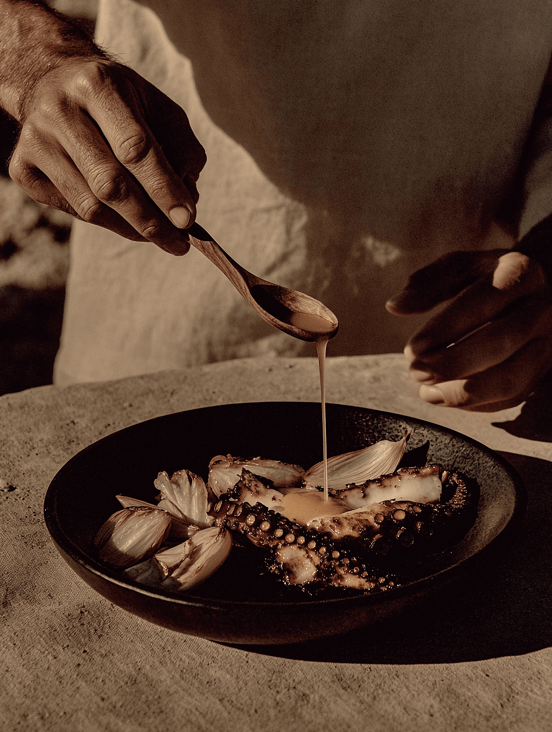 Gråhanå culinary experience — close-up of a chef drizzling sauce over roasted octopus and onions, showcasing sensory design, craftsmanship, and refined gastronomy within Astrophat Studio’s luxury hospitality branding.